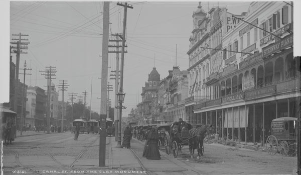 Canal Street in New Orleans seen from the Clay monument, with three animal-powered streetcars standing on the neutral ground track and cast iron galleries fronting the stores along the right side of the street.