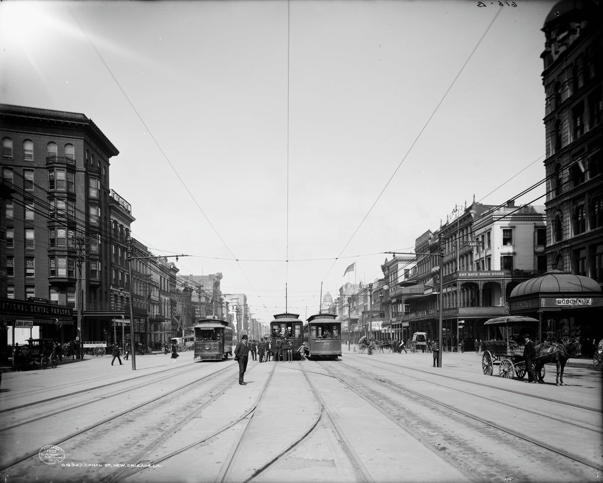 Three electric streetcars stand abreast on Canal Street in New Orleans in 1907, with pedestrians in the track and Godchaux's store under its canopy on the right.