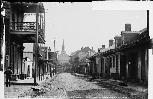 A cobbled New Orleans street restored in color, lined with iron-galleried buildings on the left and low Creole cottages on the right, closed at the far end by the trees of St. Anthony's Garden and the rear of the Cathedral of St. Louis.